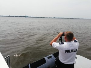 Zabezpieczenie regat żeglarskich odbywających się na Zalewie Włocławskim. Policjant obserwujący uczestników przez lornetkę. W oddali widać jachty uczestniczące w regatach