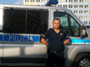 Zdjęcie pokazuje policyjny radiowóz, a przed nim sierż. sztab. Łukasza Jędrzejewskiego. Policjant przed służba pomógł kobiecie