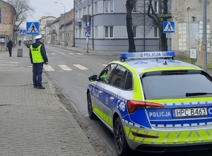 Policjant stoi przy radiowozie i patrzy na zachowania kierujących i pieszych w rejonie drogi