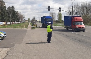 Policjant w kamizelce odblaskowej z napisem POLICJA stoi przy drodze w pobliżu przejścia dla pieszych i mierzy prędkość pojazdów